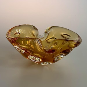 May include: An amber-coloured glass bowl in a heart shape. The bowl has a textured surface with small bubbles. The glass is a warm, golden hue, and the bowl appears to be handmade. It is likely used for decorative purposes.