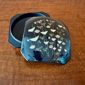 May include: Black lacquered box with a floral design. The lid features a stylized arrangement of white and green flowers. The box is open, revealing a matching black interior. The box sits on a wooden surface.