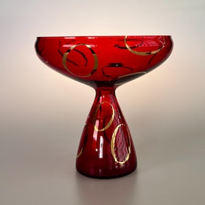 May include: A vibrant red glass bowl with a flared rim and a pedestal base. The bowl is adorned with gold circular designs. This decorative piece is suitable for serving or display.