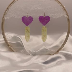 May include: A pair of dangle earrings featuring purple heart-shaped tops and light yellow, rectangular bottoms with gold flecks. The earrings hang from silver hooks. The earrings are displayed in front of a wooden embroidery hoop with white lace.