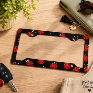 May include: Black license plate frame decorated with red ladybugs and white dots. The ladybugs have black spots. The frame is rectangular with rounded corners and holes for mounting. A matching ladybug keychain is next to the car keys.