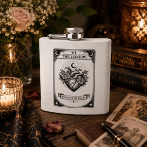 Op de afbeelding: Een witte metalen heupfles met een zwart-witte afbeelding van een tarotkaart met de titel VI THE LOVERS. Het ontwerp toont een anatomisch hart omringd door rozen en een maansikkel, met de tekst YOU HAVE MY HEART op een banner eronder.