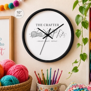 May include: A black-framed wall clock with a white face, featuring the text "THE CRAFTER" and crafting-related illustrations. The clock is 10 inches (25 cm) in diameter. The clock's design includes yarn, scissors, and needles. The phrase "CREATE - CRAFT - REPEAT" is below the illustrations.