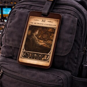 May include: A dark gray backpack with a leather luggage tag. The tag features an illustration of an air traffic controller and the text "THE AIR TRAFFIC CONTROLLER". The tag is brown with a decorative border and the Roman numeral XI.