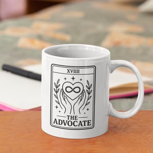 May include: White ceramic mug with a black graphic of hands holding an infinity heart, framed by leaves and stars. The text "XVIII" and "THE ADVOCATE" are also in black. The mug is on a wooden surface.