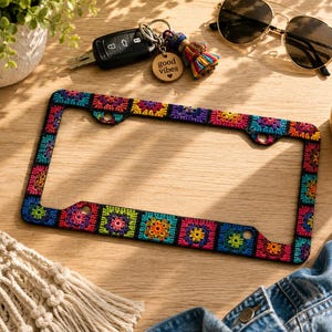 May include: A colorful license plate frame with a crochet design featuring squares in various colors like red, yellow, blue, and green. The frame is black and has mounting holes. Other items include sunglasses, keys, and a drink.