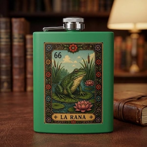 May include: A matte green metal flask featuring a rectangular graphic print of a frog on a lily pad in a pond. The print includes the number 66, the text LA RANA, and a decorative floral border. The flask has a silver screw-top cap at the centre.