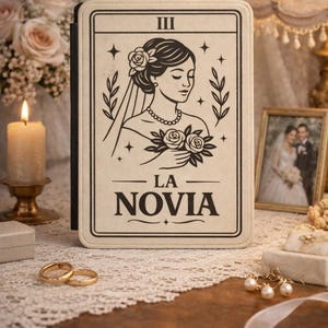 May include: A cream-colored tarot card with a black illustration of a bride, the Roman numeral III, and the words "LA NOVIA." The card is surrounded by floral and star motifs. The image is set on a wooden surface with wedding-themed accessories.