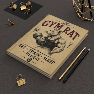 May include: A tan hardcover journal featuring a graphic of a muscular rat lifting a dumbbell. The text reads The GYM RAT, 24, 7, and EAT • TRAIN • SLEEP • REPEAT. The cover includes a small kettlebell icon and a star. The journal is placed on a dark surface.