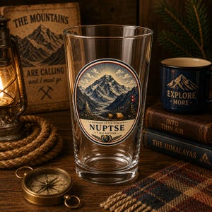 Puede incluir: Un vaso de cristal transparente con un gráfico circular del pico Nuptse. El diseño incluye el texto WONDERS OF THE WORLD, NUPTSE y 7.861 METROS, junto con una ilustración de una montaña nevada, banderas de oración y tiendas de campaña.