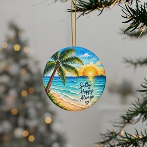 May include: A round ceramic ornament featuring a vibrant watercolor beach scene with a palm tree, sandy shore, blue ocean, and a golden sunset. The text "Salty Happy Always" is printed in black script above a small heart symbol on the water.