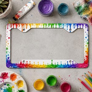 Könnte beinhalten: Ein rechteckiger Kennzeichenhalter mit einem lebendigen Regenbogen-Farbtropfenmuster. Das Design zeigt ein Farbspektrum mit Rot, Orange, Gelb, Grün, Blau und Violett, wobei Farbspritzer und Tropfen über den weißen Rahmen des Halters verlaufen.