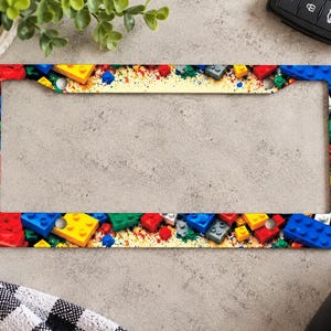 Könnte beinhalten: Ein rechteckiger Kennzeichenhalter mit einem bunten Muster aus ineinandergreifenden Kunststoff-Bausteinen in Rot, Blau, Gelb und Grün, vor einem hellen Hintergrund mit farbigen Sprenkeln und vier Befestigungslöchern für die Montage.