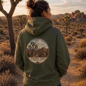 Pode incluir: Um moletom verde-oliva com um gráfico circular de uma paisagem do Parque Nacional Joshua Tree nas costas. O moletom tem um capuz com cordão e é usado em um ambiente desértico.