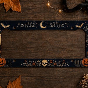May include: A rectangular black license plate frame featuring a decorative Halloween theme. The design includes a central crescent moon, a skull, two jack-o'-lanterns, a ghost, bats, and floral motifs printed in orange, white, and gold tones.