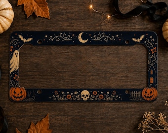 May include: A rectangular black number plate frame featuring a decorative Halloween theme. The design includes a central crescent moon, a skull, two jack-o'-lanterns, a ghost, bats, and floral motifs printed in orange, white, and gold tones.