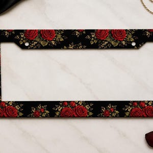 May include: A rectangular license plate frame featuring a black background decorated with a repeating pattern of deep red roses and green leaves. The frame has two circular mounting holes at the top and a wide border design throughout the piece.