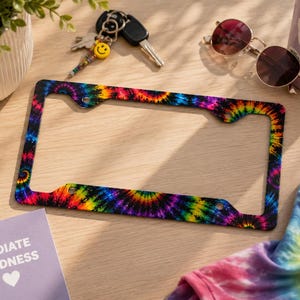 May include: A license plate frame with a vibrant tie-dye pattern in shades of blue, purple, yellow, and orange. The frame is rectangular with rounded corners and mounting holes. A small card with the words "RADIATE KINDNESS" and a heart is visible.