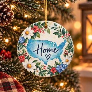 May include: A round ceramic ornament featuring a watercolour map of the United States in blue, centred with the word Home and a small heart symbol. The map is surrounded by a decorative border of white daisies, red flowers, and green leaves.