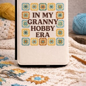 May include: A cream-colored case with the text "IN MY GRANNY HOBBY ERA" and colorful granny square designs. The case is surrounded by yarn, crochet projects, and a pair of scissors, suggesting a crafting theme.