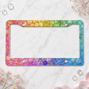 May include: A rectangular license plate frame featuring a vibrant rainbow gradient pattern composed of numerous circular, glittery, and textured dots. The frame has four mounting holes and a smooth, flat surface with a colorful, multi-hued design.
