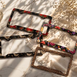 May include: Four license plate frames with various designs. One features red roses on a black background, another has butterflies, a third showcases colorful flowers, and the last has a leopard print pattern. The frames are rectangular with rounded corners.
