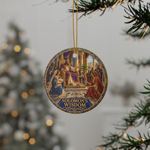 Può includere: Un ornamento rotondo in ceramica con un'illustrazione dettagliata di Re Salomone seduto su un trono, circondato da figure in abiti. Il testo "SOLOMON'S WISDOM" è stampato in oro nella parte inferiore. L'ornamento pende da un filo dorato.