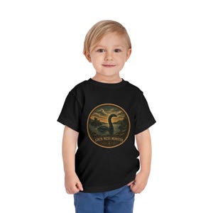 May include: A black short-sleeve t-shirt featuring a circular graphic on the chest. The design depicts a lake scene with the Loch Ness Monster, a castle in the distance, and clouds, with the text LOCH NESS MONSTER printed at the bottom.