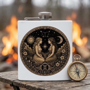 May include: A white metal flask featuring a circular illustration of the Gemini zodiac sign with two figures, a sun, a moon, and stars. The flask has a silver screw-top lid and is positioned next to a round brass compass on a wooden surface.