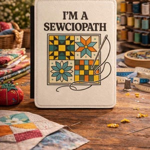 May include: A cream-colored notebook with the text "I'M A SEWCIOPATH" and a colorful patchwork design. The design includes a needle and thread. The notebook is on a wooden table with sewing supplies.