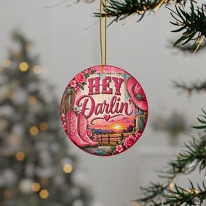 May include: A round ceramic ornament hanging by a gold string, featuring a sunset landscape with pink cowboy boots, a pink cowboy hat, and floral accents. The text "HEY Darlin'" is printed in pink lettering across the center of the design.