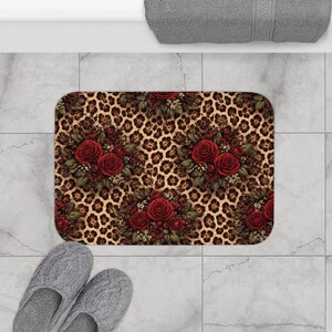 May include: Rectangular bath mat featuring a leopard print background with clusters of red roses and greenery. The mat has rounded corners and is placed on a tiled floor. Gray slippers are visible.