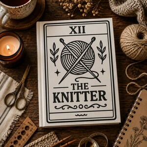 May include: A white hardcover book with black line art featuring a ball of yarn crossed by two knitting needles, the Roman numeral XII, and the text THE KNITTER. The book is surrounded by a cup of tea, a candle, scissors, a ruler, and yarn.