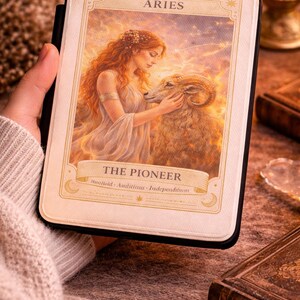 May include: A tarot card featuring the Aries zodiac sign, with an illustration of a woman and a ram. The card is labeled "The Pioneer" with the words "Bold, Ambitious, Independent" below. The card is held in a black case.