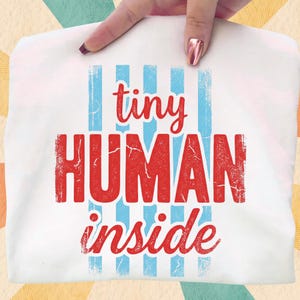 May include: White t-shirt with the words "tiny HUMAN inside" in red and blue. The text is distressed, giving it a vintage look. Blue vertical stripes are behind the word "tiny".