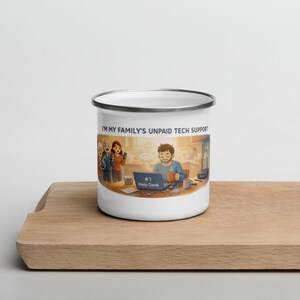 Op de afbeelding: Witte emaille mok met een zwarte rand. De mok heeft een illustratie van een persoon die op een laptop werkt met de tekst "I'M MY FAMILY'S UNPAID TECH SUPPORT" en "#1 Help Desk". De mok staat op een houten oppervlak.