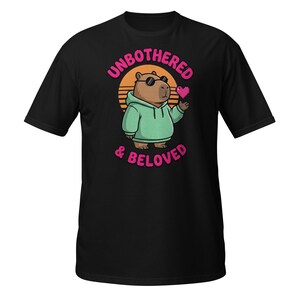 Könnte beinhalten: Schwarzes T-Shirt mit einem Cartoon-Wasserschwein, das eine Sonnenbrille und einen grünen Kapuzenpullover trägt und ein pixeliges rosa Herz hält. Der Text "UNBOTHERED & BELOVED" ist in Rosa über und unter dem Wasserschwein aufgedruckt, mit einer Sonnenuntergangsgrafik im Hintergrund.