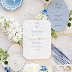 May include: Elegant wedding invitation suite with embossed details, featuring a blue plate with a bird design, white and blue flowers, and a ring box with rings. The suite includes invitation cards and envelopes.