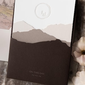 May include: A wedding invitation with a layered mountain design. The invitation features a white top with a circular embossed monogram and a landscape of torn paper in shades of gray and brown. The bottom is a dark brown pocket with the text "ON THIS DAY 03.05.24".