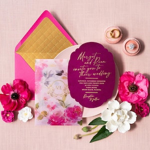 May include: A wedding invitation suite featuring a magenta invitation with gold script, a floral envelope, and a floral card. Two ring boxes and pink and white flowers complete the arrangement on a pink background.