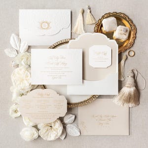May include: An elegant wedding invitation suite in ivory and gold. The set includes invitation cards, envelopes, and a decorative tray with ring boxes. Floral accents and tassels add to the sophisticated design.