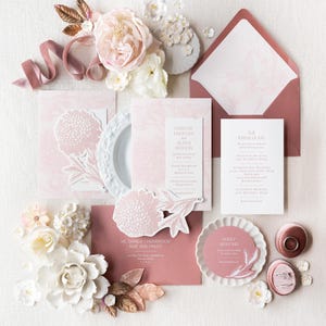 May include: A flat lay of wedding stationery in shades of pink and white. The arrangement includes invitations, envelopes, and decorative floral elements. The text on the invitations includes names and event details. A ribbon and decorative flowers complete the design.