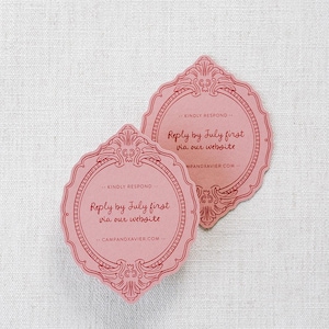 May include: Two pink, ornate invitation cards with a decorative frame design. The cards feature the text "Reply by July first via our website" and the website address. The cards are on a white background.