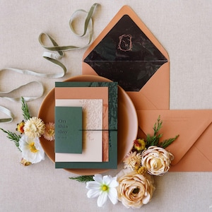 May include: A styled wedding invitation suite featuring a terracotta envelope, a dark green invitation card with the text "On this day", and a matching plate. The arrangement includes flowers and a velvet ribbon.