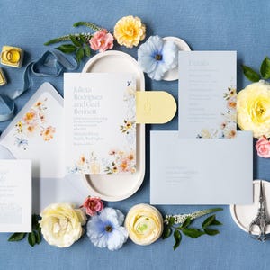 May include: Wedding invitation suite with floral accents, including invitation, reply card, details card, and envelope. The suite features the names "Julieta Rodriguez and Gael Bennett". The design includes yellow, pink, and blue flowers, with a blue ribbon and two ring boxes.