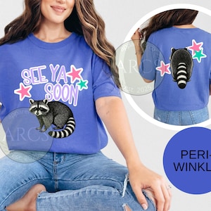 May include: Periwinkle blue t-shirt with a raccoon graphic and the text "See Ya Soon". The design includes pink and green stars. The back of the shirt also features a raccoon and stars.