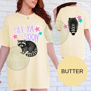 May include: Butter-colored t-shirt with a raccoon graphic and the text "SEE YA SOON". The front of the shirt features a raccoon illustration with stars. The back of the shirt also has a raccoon graphic and stars.