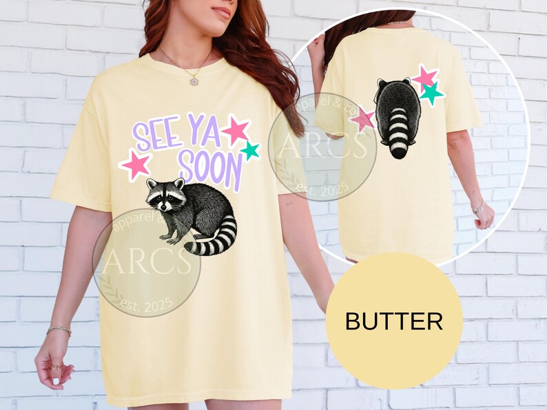 May include: Butter-colored t-shirt with a raccoon graphic and the text "SEE YA SOON". The front of the shirt features a raccoon illustration with stars. The back of the shirt also has a raccoon graphic and stars.