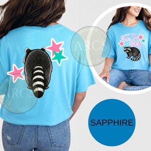 May include: Light blue t-shirt featuring a raccoon graphic with a striped tail and pink and green star accents. The shirt also has the text "See Ya Soon" in pink and green. The shirt is a sapphire color.