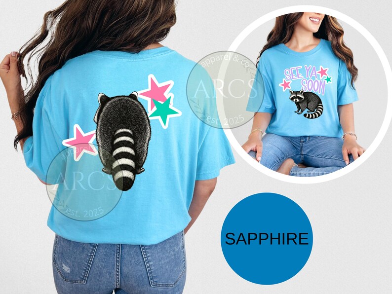 May include: Light blue t-shirt featuring a raccoon graphic with a striped tail and pink and green star accents. The shirt also has the text "See Ya Soon" in pink and green. The shirt is a sapphire color.